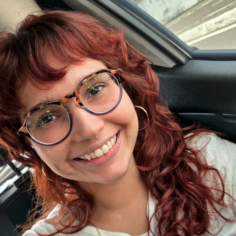 Bárbara Annitza sorrindo, de óculos de armação tartaruga e cabelo ruivo cacheado